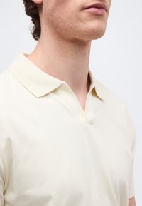 Νεαρό άτομο που φοράει πουκάμισο κρεμ με γιακά και άνοιγμα σε σχήμα V, μπροστά σε απλό ανοιχτό φόντο.