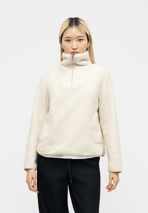 Jeune femme portant un pull polaire crème avec un col zippé haut et un pantalon noir, debout devant un fond blanc uni.