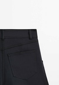 Vue arrière d'un pantalon noir montrant la ceinture, les passants de ceinture et une poche arrière cousue unique sur fond blanc uni.