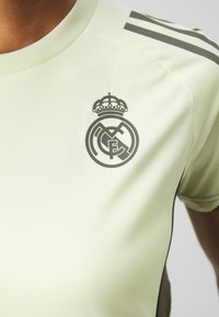 Maglietta sportiva verde chiaro a maniche corte, con un logo Real Madrid grigio scuro e dettagli sul collo e sulle spalle.