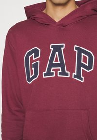 Personne portant un sweat à capuche bordeaux avec de grandes lettres "GAP" en bleu marine et blanc sur la poitrine, ainsi qu'une poche avant kangourou.