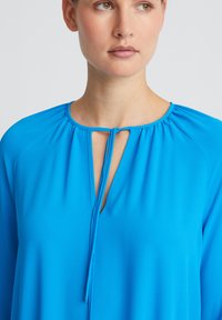 Blauwe blouse met een ronde halslijn, keyhole-detail en striksluiting. Gemaakt van gladde, lichte stof, met lange mouwen en subtiele plooien.