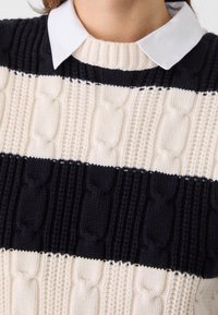 Pull en tricot présentant des rayures horizontales crème et marine, avec des motifs en câble texturé. Le col blanc ajoute un contraste.
