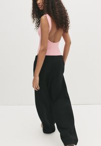 Vrouw met krullend haar die loopt, gekleed in een mouwloze roze top met een lage rug en losse zwarte broek tegen een effen achtergrond.