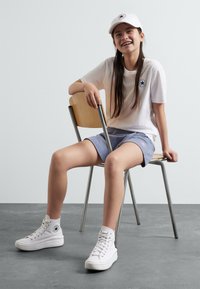 T-shirt blanc avec un logo, shorts gris clair et baskets montantes blanches. Assis sur une chaise en métal devant un fond uni.