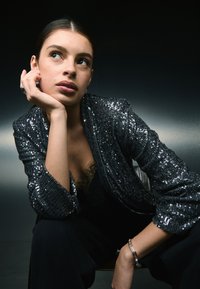 Čierna flitrovaná bunda vo voľnom strihu, kombinovaná s čiernym čipkovaným topom. Pozadie je tmavé, čo zvýrazňuje trblietavú textúru bundy.