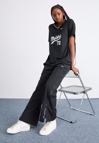 Črna majica Nike z belim logotipom in tiskom "72", v kombinaciji s črnimi hlačami z zadrgami. Bela športna obutev Nike zaokroži videz.