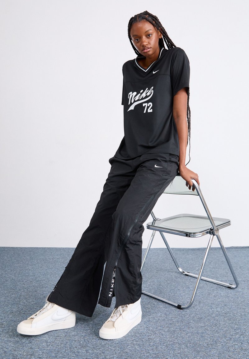 T-shirt noir Nike avec logo blanc et impression « 72 », associé à un pantalon noir avec des accents zippés. Des baskets blanches Nike complètent le look.