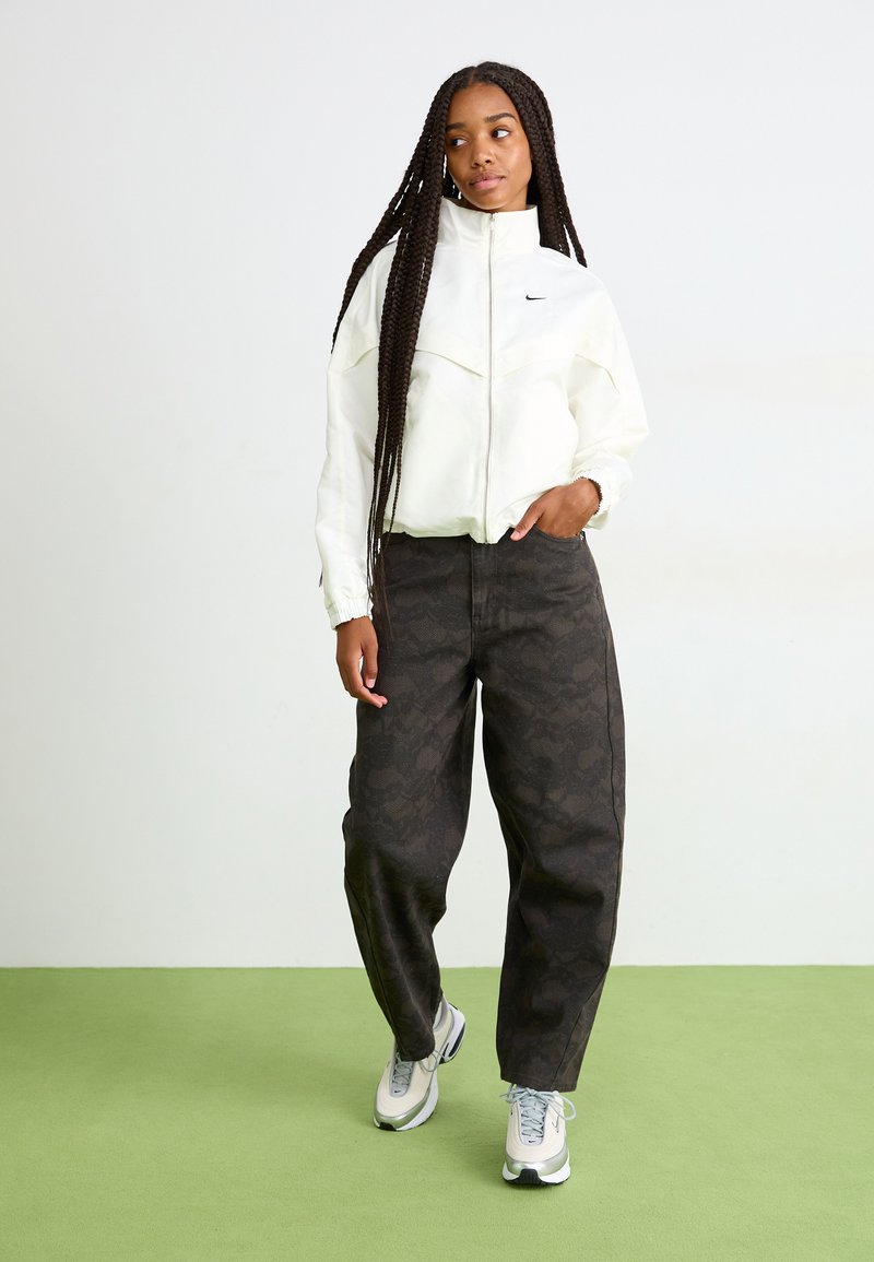 Woman with long braids wearing white zip-up jacket, dark patterned wide pants, and white sneakers standing on green floor against white wall.