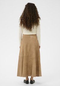 Tumši zamšs maxi svārki ar izplestu formu, ar aizmugures rāvējslēdzēju un gludu tekstūru, kombinēti ar krēmkrāsas ribotu džemperi un melnām iešļūcenēm.