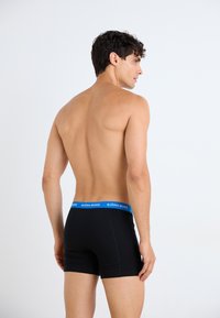 Schwarze Boxershorts mit einem blauen Bund, der das Logo "BJÖRN BORG" trägt. Das Material wirkt glatt, das enge Design betont den Oberschenkel.
