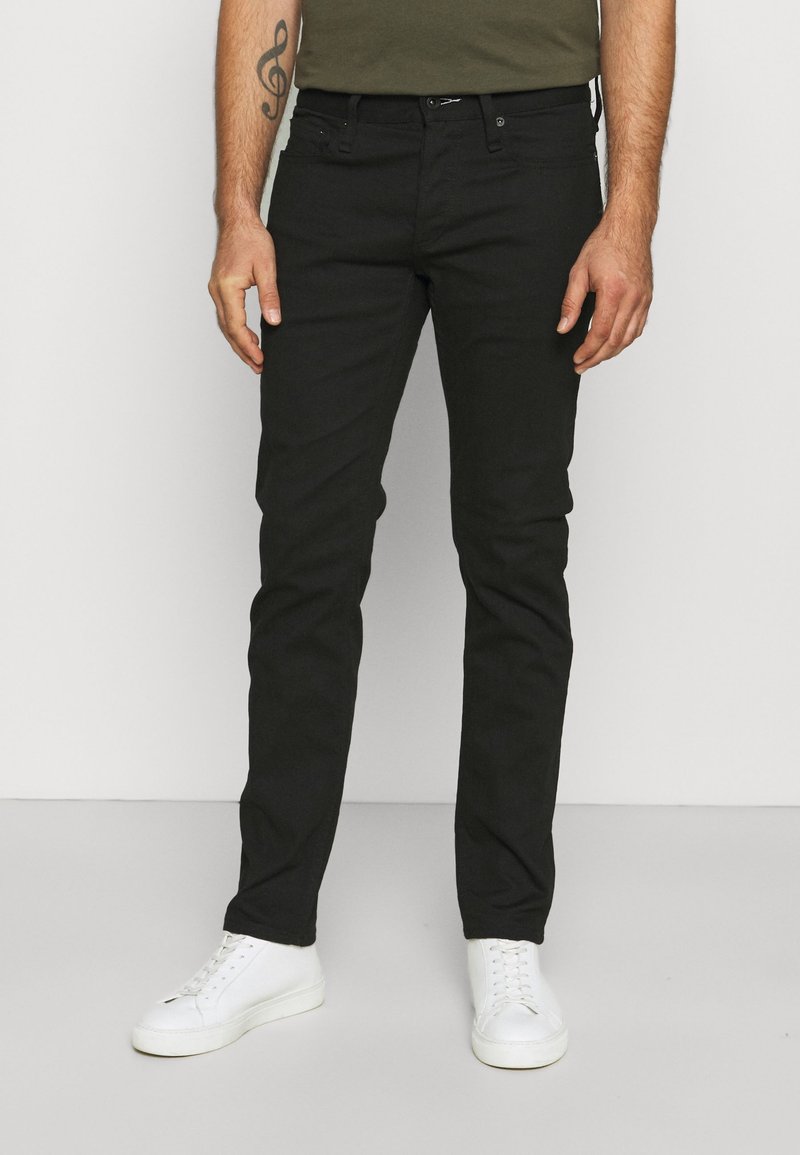 Jeans in denim nero con una vestibilità slim, dotati di cinque tasche, passanti per cintura e chiusura classica con bottone e zip. Indossati con sneakers bianche.