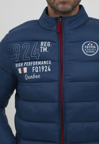 Tamsiai mėlynas pūkinis švarkas su aukštu apykakle, raudona užtrauktuku ir baltais spaudiniais, įskaitant "924 REG." ir "Aukštos kokybės", taip pat logotipo detales.