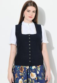 Marineblauw corduroy vest met een vierkante halslijn, sluiting met zilveren knopen, gedragen over een witte geplooide blouse met kanten afwerking. Onder een rok met bloemmotief.