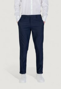 Homme portant un pantalon habillé bleu marine, une chemise blanche à boutons, et des baskets blanches, se tenant avec les mains dans les poches sur un fond uni.