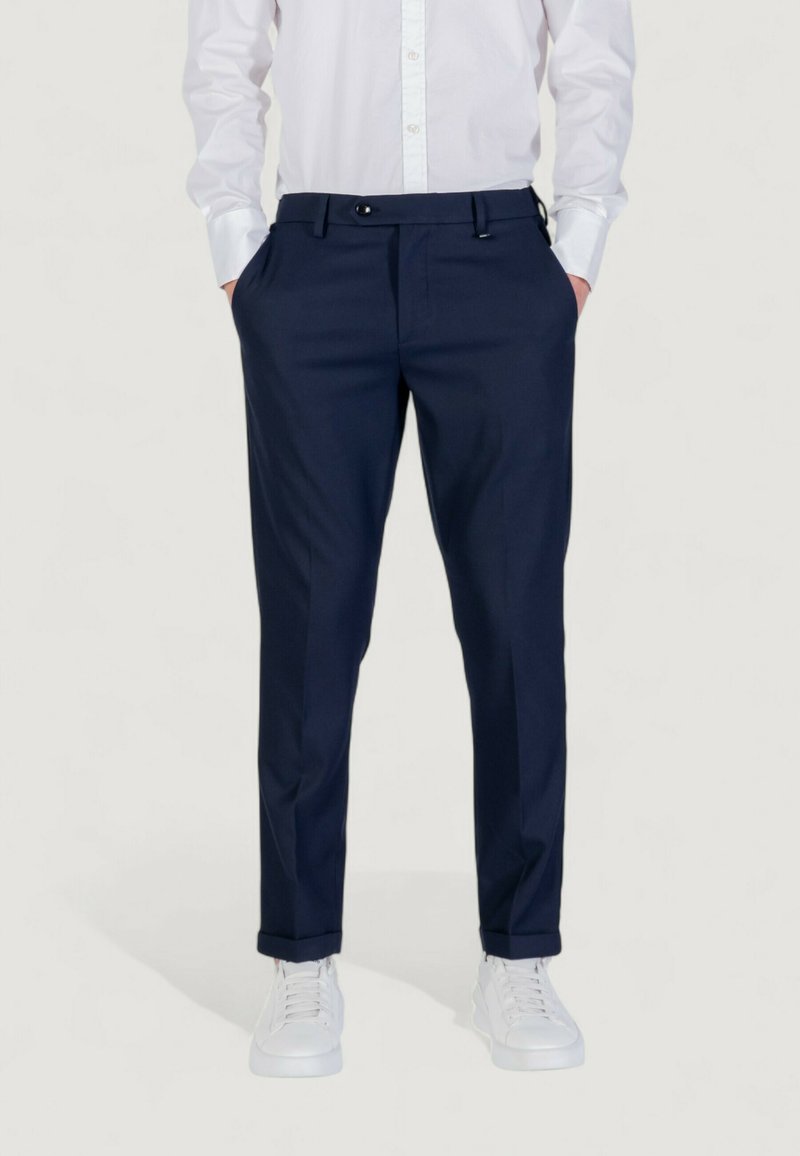 Homme portant un pantalon habillé bleu marine, une chemise blanche à boutons, et des baskets blanches, se tenant avec les mains dans les poches sur un fond uni.