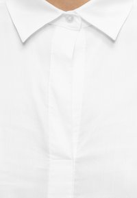 Gros plan d'une personne portant une chemise blanche à col boutonnée en haut, montrant les détails du col et de l'encolure.