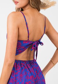 Blaues Blumen-Crop-Top mit dünnen Trägern, offener Rückseite mit Knotenverschluss und detailliertem roten Blumenmuster. Weicher Stoff mit leichtem Glanz.