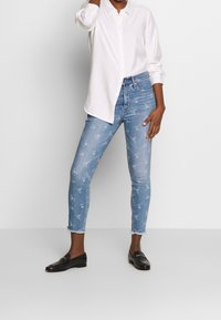 Camisa blanca de manga larga, vaqueros de denim azul claro con estampado floral. Los vaqueros tienen un largo recortado y los dobladillos deshilachados. Mocasines negros.
