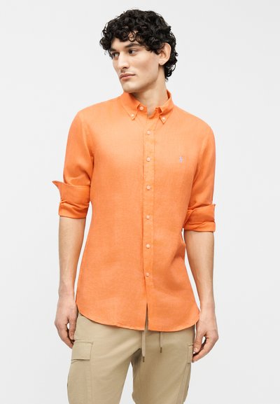 Άνδρας με σγουρά μαλλιά φορώντας πορτοκαλί πουκάμισο με κουμπιά και μανίκια γυρισμένα, καθώς και μπεζ παντελόνι cargo, κοιτάζει στο πλάι μπροστά σε μονόχρωμο φόντο.