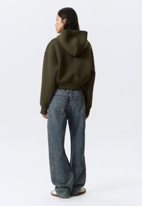 Γυναίκα που στέκεται με την πλάτη, φοράει μια κοντή φούτερ με κουκούλα σε ελιά πράσινο, φαρδιές μπλε τζιν και λευκά αθλητικά, μπροστά σε ένα μονόχρωμο φόντο.