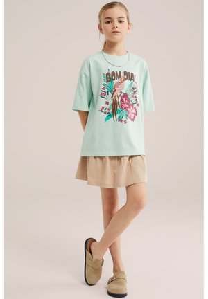 Girl wearing mint green oversized T-shirt with bird and flower print, beige skirt, and beige slip-on shoes standing with crossed legs.