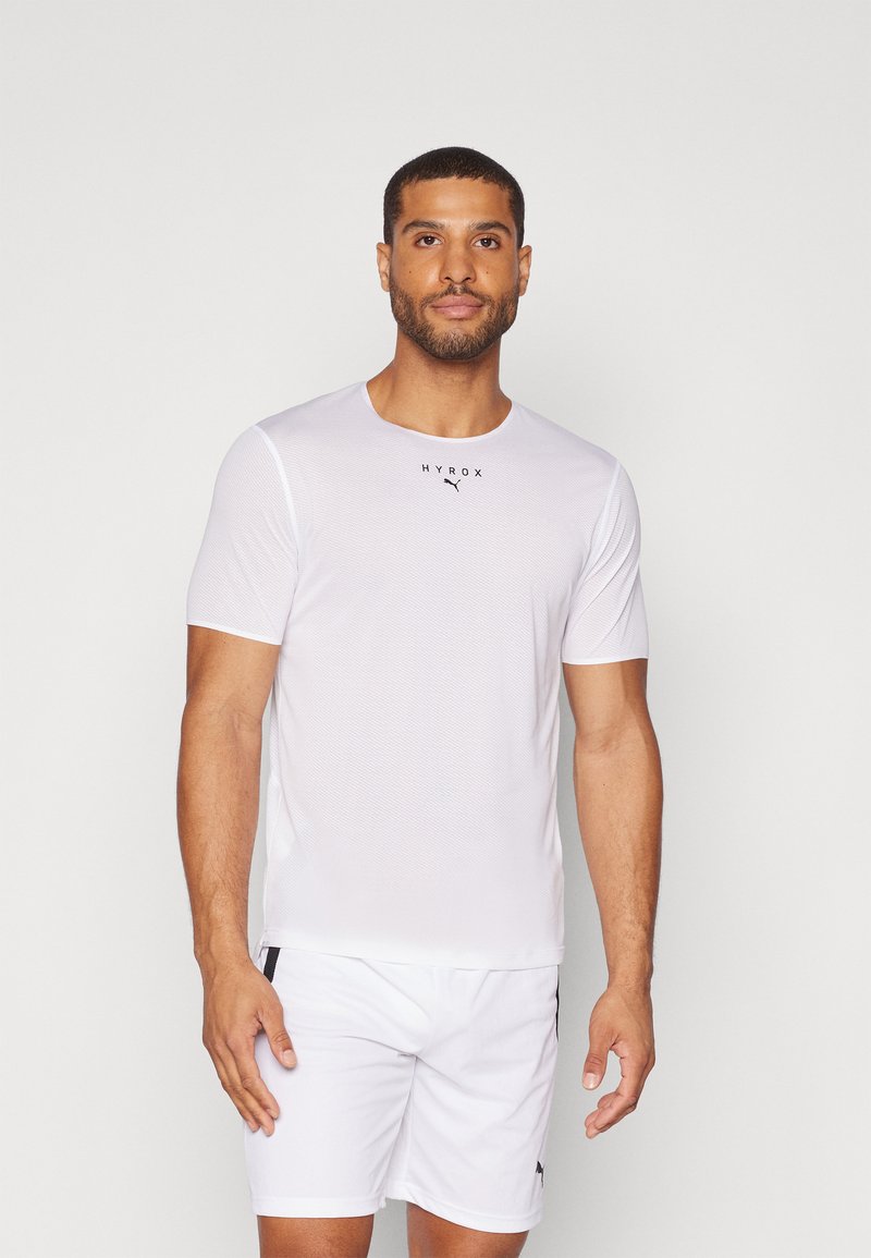 T-shirt de sport blanc à manches courtes, col rond et tissu texturé ; présente un logo noir et un branding subtil sur la poitrine.