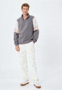 Grijze sweater met ritssluiting en crèmekleurige mouwen, gecombineerd met witte cargobroek en beige sneakers, tegen een effen lichte achtergrond.