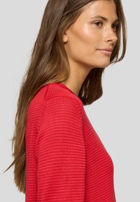 Frau mit langen braunen Haaren, die einen roten Strickpullover trägt, im Profil vor einem neutralen Hintergrund dargestellt.