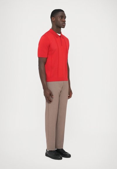 Punainen neulottu poolopaita, jossa on lyhyet hihat ja tekstuuriset paneelit; yhdistetty beigenvärisiin räätälöityihin housuihin ja mustiin tennareihin.