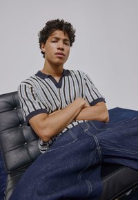 Polo à manches courtes rayé avec motifs bleus, crème et noirs ; jean en denim foncé ; assis sur une chaise en cuir noir.