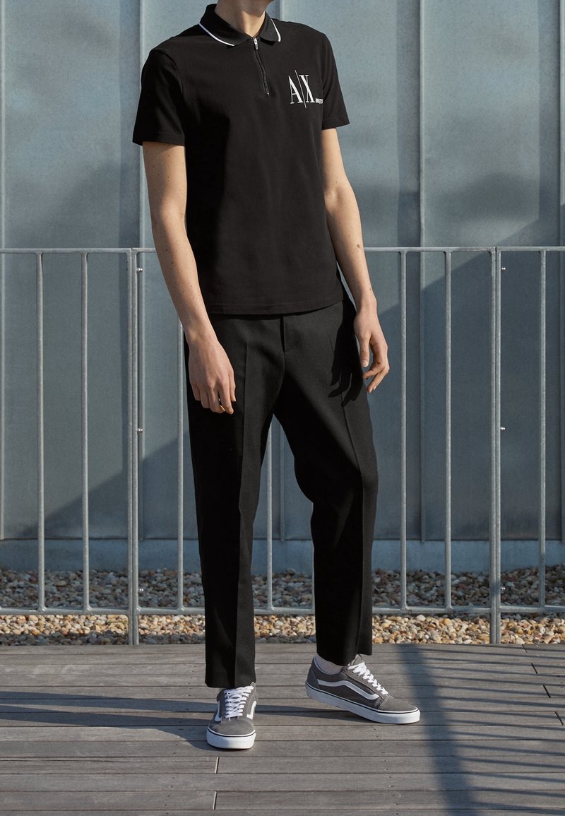 Homme portant un polo noir à manches courtes avec un logo blanc, un pantalon noir et des baskets grises, debout sur un sol en bois près d'une rambarde en métal.