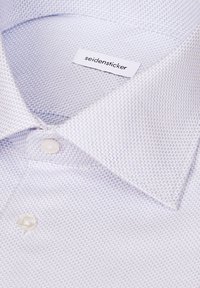 Gros plan d'une chemise blanche avec un fin motif zigzag bleu, des boutons beige et une étiquette indiquant le nom de la marque "seidensticker."