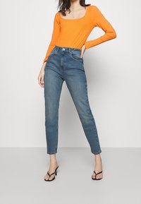 Frau mit einem figurbetonten leuchtend orangefarbenen Langarmtop, hochgeschnittenen blauen Jeans und schwarzen Absatzsandalen, die vor einem schlichten Hintergrund steht.