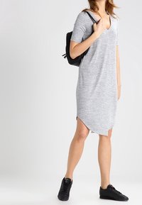 Abito grigio a maglia con scollatura a V e maniche corte, caratterizzato da un orlo leggermente asimmetrico. Indossato con sneaker nere e uno zaino.