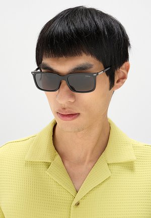 Uomo che indossa occhiali da sole rettangolari neri e una camicia verde chiaro a trama con colletto, guarda leggermente verso il basso e di lato.
