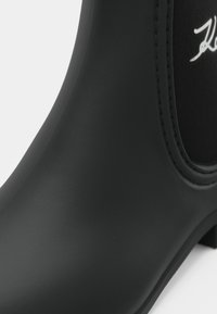 Bota de tornozelo preta em borracha com um acabamento suave, bico arredondado e detalhes costurados. Apresenta um pequeno logótipo na lateral.