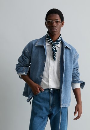 Man die doorzichtige bril, spijkerjack en spijkerbroek draagt, een gestreept shirt en een donkere gestreepte neksjaal, staand tegen een effen grijze achtergrond.