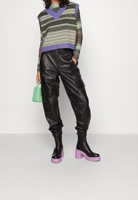 Schwarze Leder-Cargohose; mehrfarbiges, ärmelloses Strickoberteil mit Diamantmuster; klobige schwarze und violette Stiefel mit Absatz; kleine grüne Handtasche.