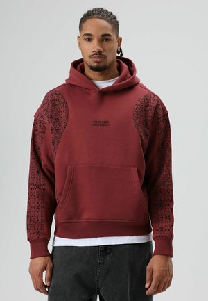 Bordeauxrode hoodie met zwarte mouwen met patroon en een print aan de voorkant. Voorzien van een grote capuchon en een buidelzak. Zacht katoenen materiaal, comfortabele pasvorm.