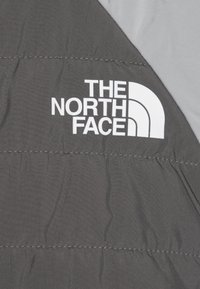 Šedá zateplená bunda s prošívanými části a bílým logem "THE NORTH FACE" na levém horním rohu. Hladký povrch, lehký materiál.