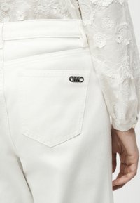 Τσέπη λευκού τζιν με ψηλή μέση και ασημένια καρφίτσα με το λογότυπο MK, συνδυασμένη με διάφανη λευκή μπλούζα με κεντήματα λουλουδιών στο μανίκι.