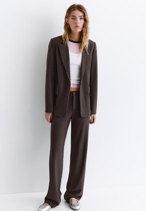 Donna con capelli biondi mossi, indossa un completo marrone scuro con pantaloni a gamba larga, un top bianco corto e scarpe piatte argento, in piedi contro un muro bianco.