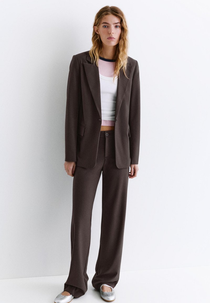 Femme aux cheveux blonds ondulés portant un tailleur marron foncé avec un pantalon à jambes larges, un crop top blanc et des chaussures plates argentées, debout contre un mur blanc.
