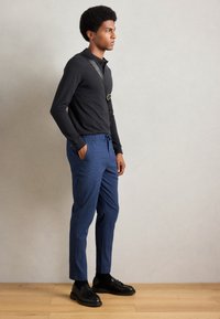 Zwarte lange mouwen shirt met een gestructureerde kraag, blauwe getextureerde broek met een trekkoord in de taille, en zwarte instappers op een houten vloer.