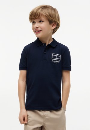 Lyshåret dreng smiler, iført en mørkeblå polo skjorte med logo på brystet og beige shorts, står foran en hvid baggrund.