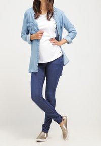 Kvinna med en ljusblå denimskjorta över en vit t-shirt, mörkblå jeans och metalliska slip-on sneakers, stående med korsade ben.