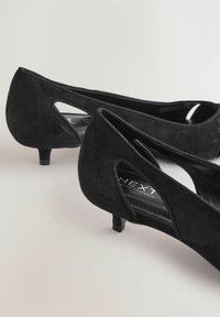 Par crnih cipela od brušene kože s niskom petom i izrezima sa strane na svijetloj površini, prikazanih pod kutom kako bi se vidjeli prednji i stražnji dizajni.