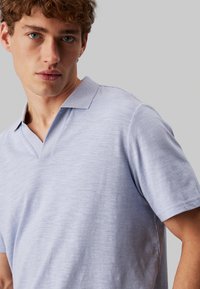 Polo de color lavanda claro con cuello en V y mangas cortas, confeccionado en una tela texturizada, que presenta un pequeño logo en el pecho.