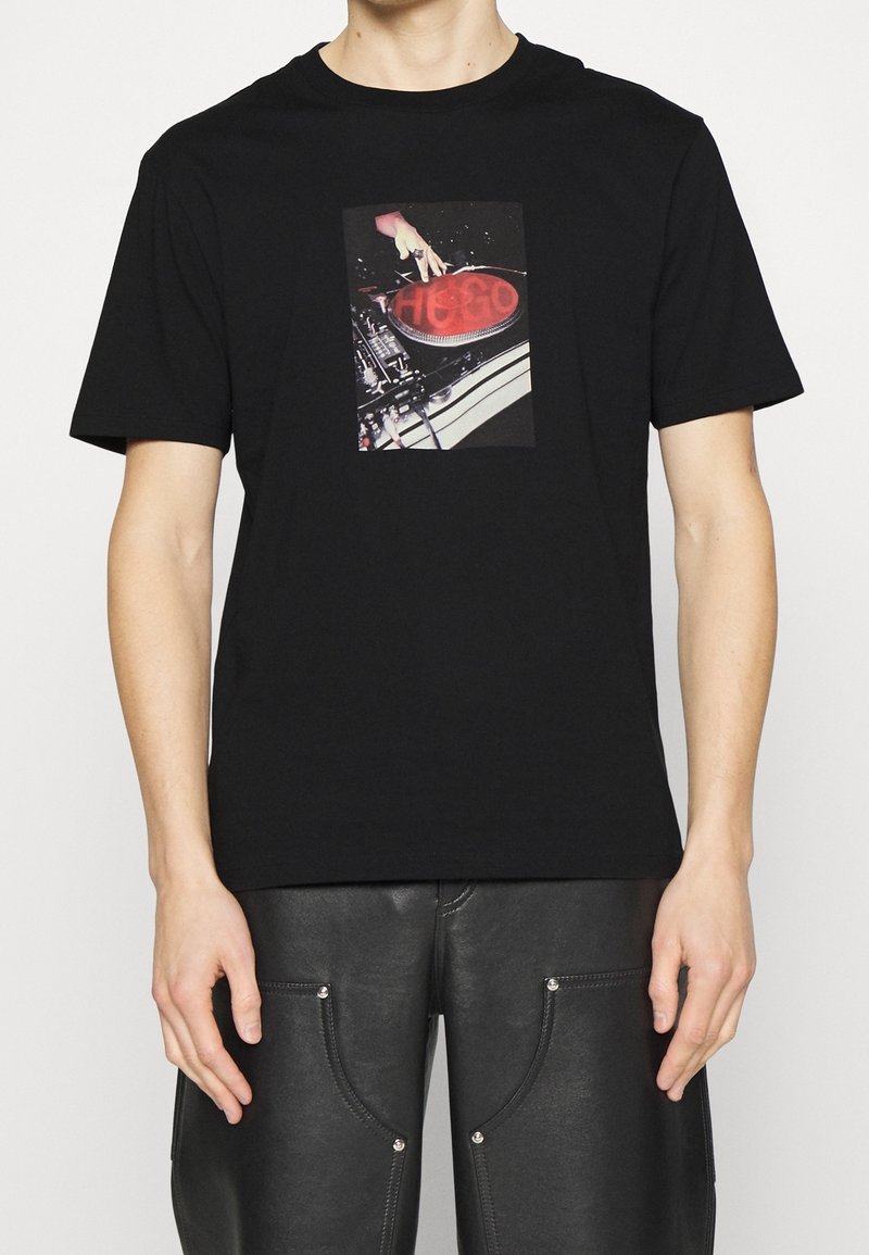 T-shirt en coton noir avec un graphique central représentant une main sur une platine, accompagné de texte rouge et blanc. Manches courtes et coupe standard.
